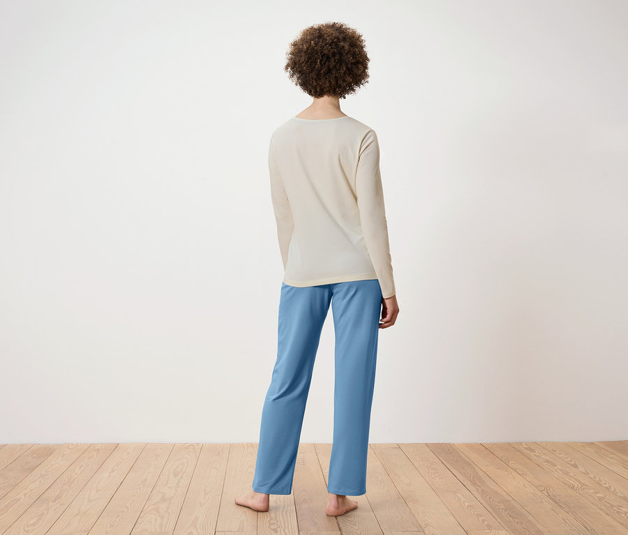 De dos, une femme aux cheveux bouclés porte un haut beige à manches longues et un pantalon bleu. Elle se tient debout sur un plancher en bois.