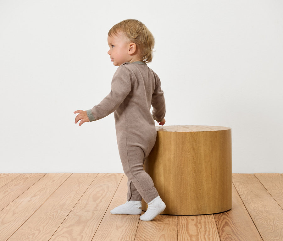 Un bébé en combinaison marron s'appuie sur une table d'appoint en bois.