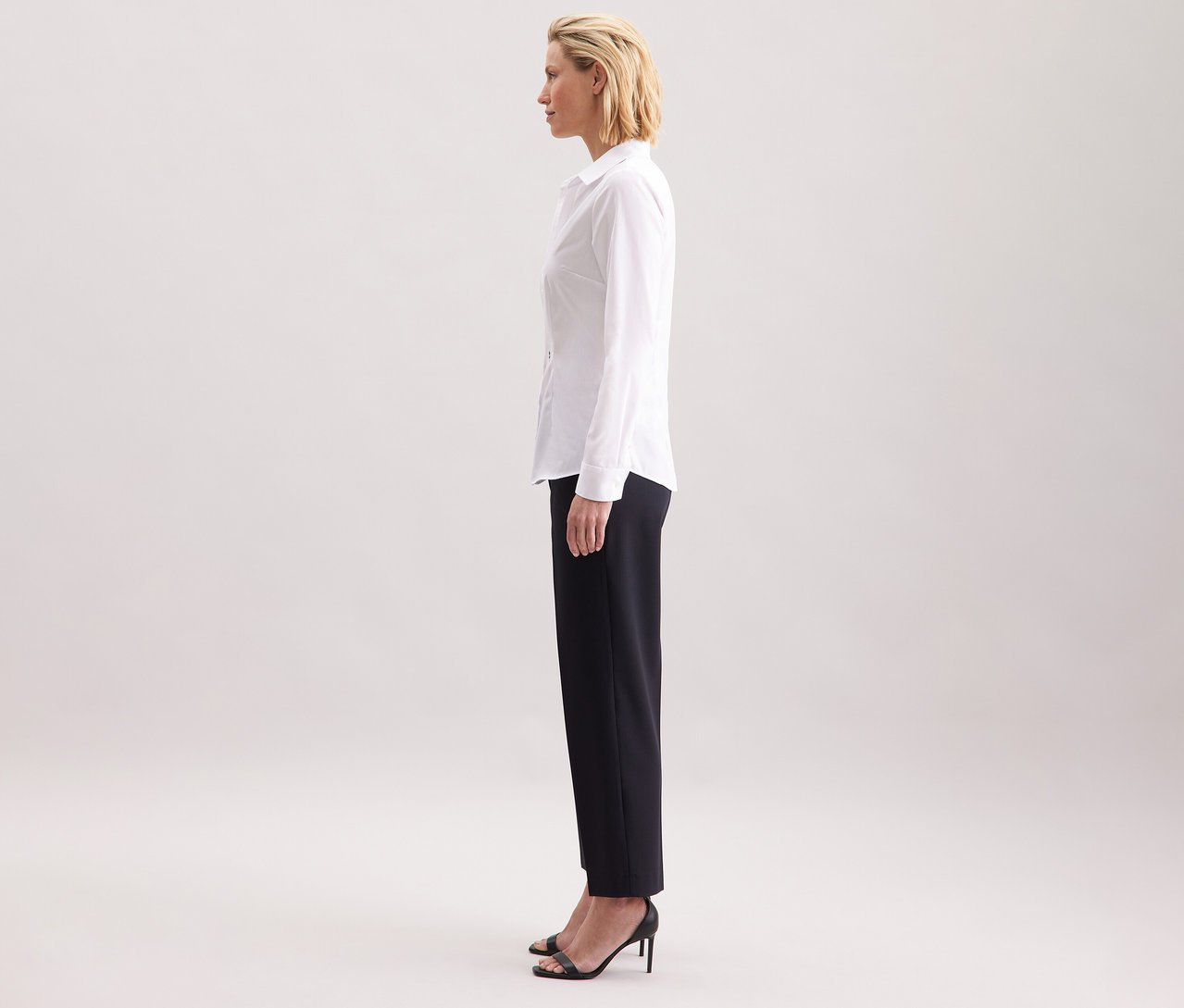 Une femme de profil portant une chemise blanche Slim Fit non iron Seidensticker, associée à un pantalon foncé et des talons hauts noirs.