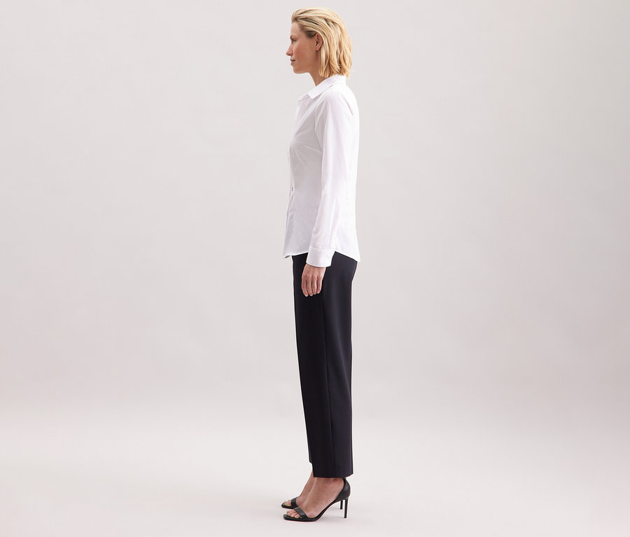 Une femme de profil portant une chemise blanche Slim Fit non iron Seidensticker, associée à un pantalon foncé et des talons hauts noirs.