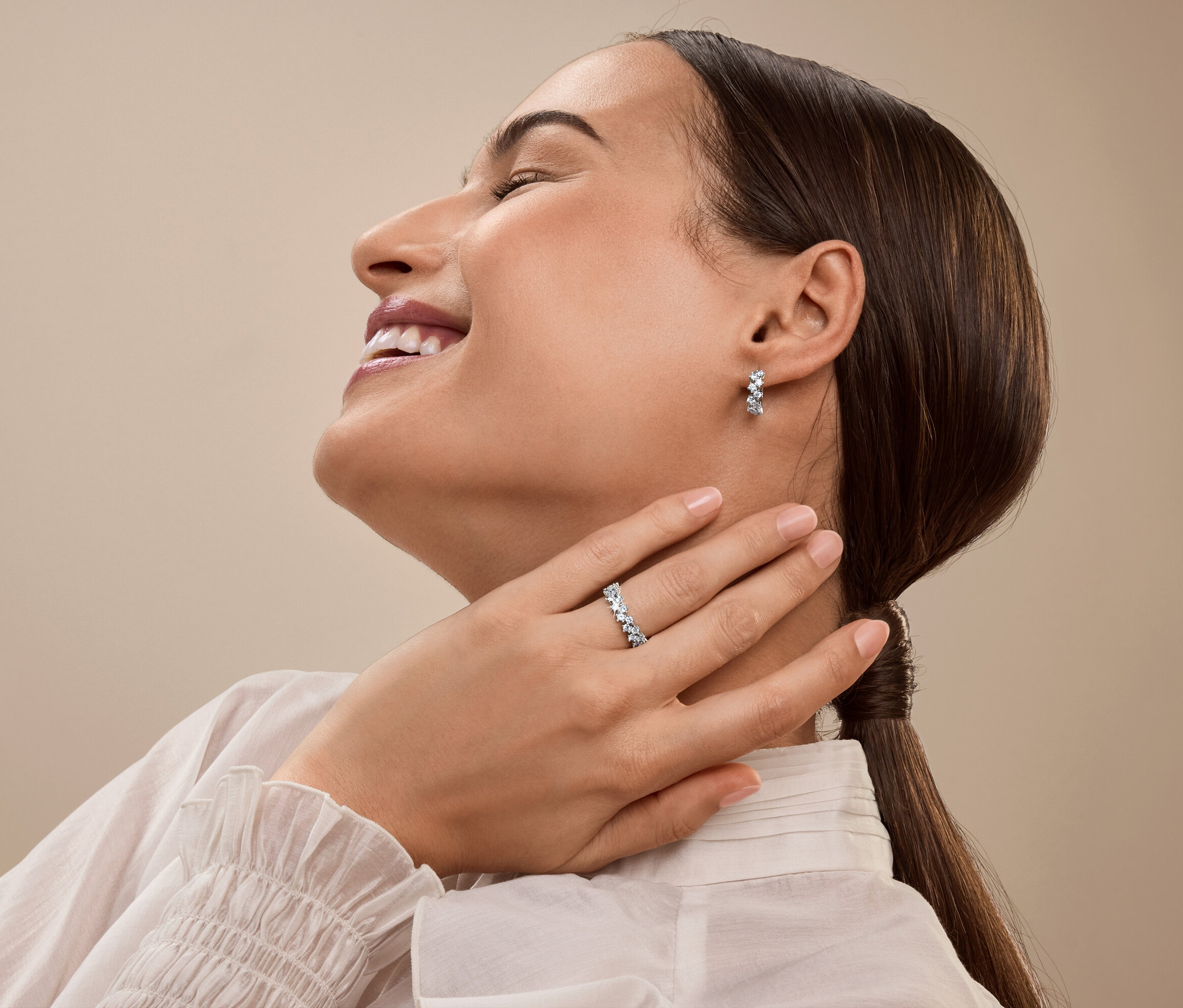 Femme souriante avec une bague en argent et des boucles d'oreilles avec des pierres scintillantes se touche le cou.
