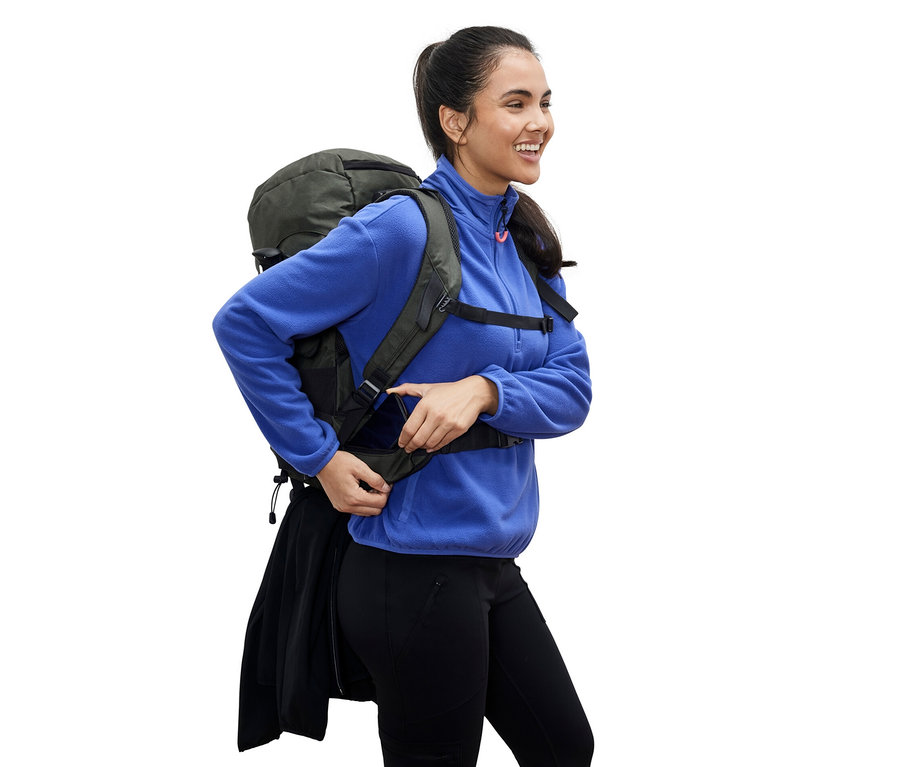 Une femme souriante vêtue d'une veste en polaire bleue ajuste la sangle d'un sac à dos de randonnée vert de 40
ol.