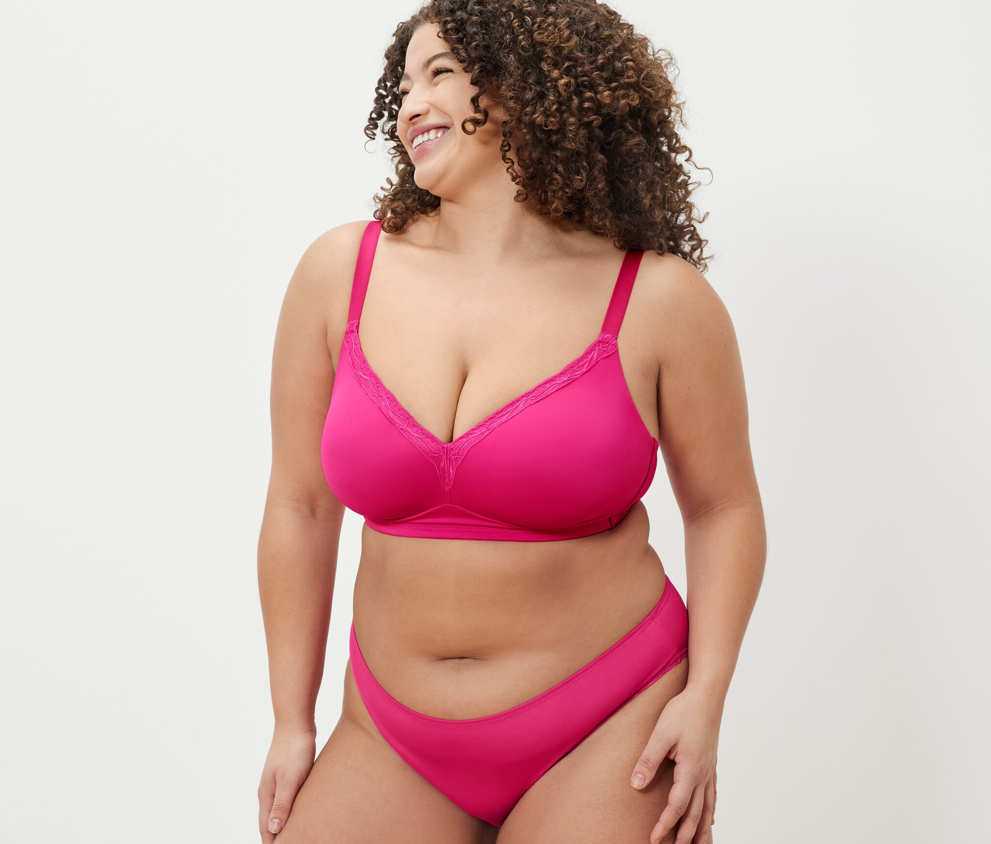 Femme aux cheveux bouclés portant soutien-gorge souple et culotte roses, regardant par-dessus son épaule et souriant.