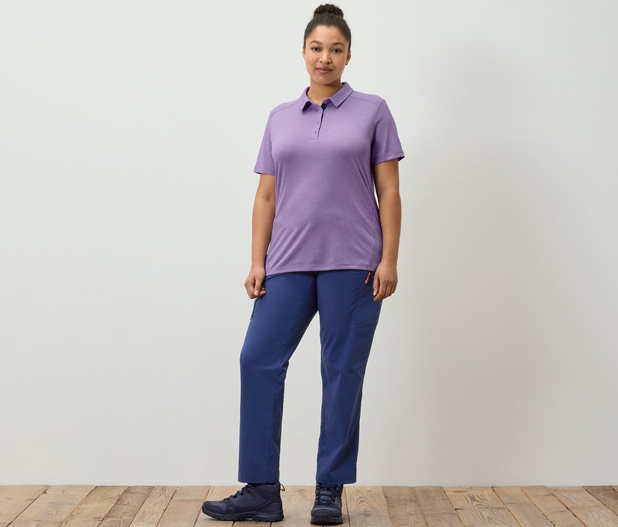 Une femme pose sur un plancher en bois en pantalon d'extérieur bleu foncé et polo fonctionnel violet.