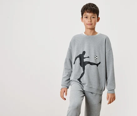 Un garçon porte un sweat-shirt gris avec une silhouette noire d'un joueur de football et un ballon dessus, ainsi qu'un pantalon de jogging gris.