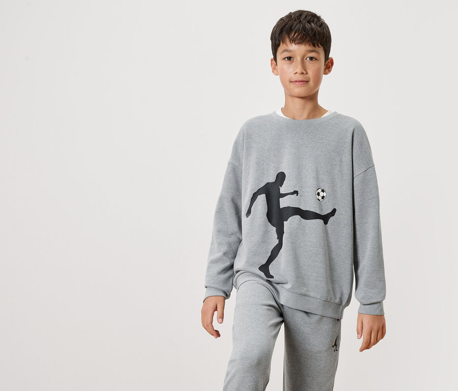 Un garçon porte un sweat-shirt gris avec une silhouette noire d'un joueur de football et un ballon dessus, ainsi qu'un pantalon de jogging gris.