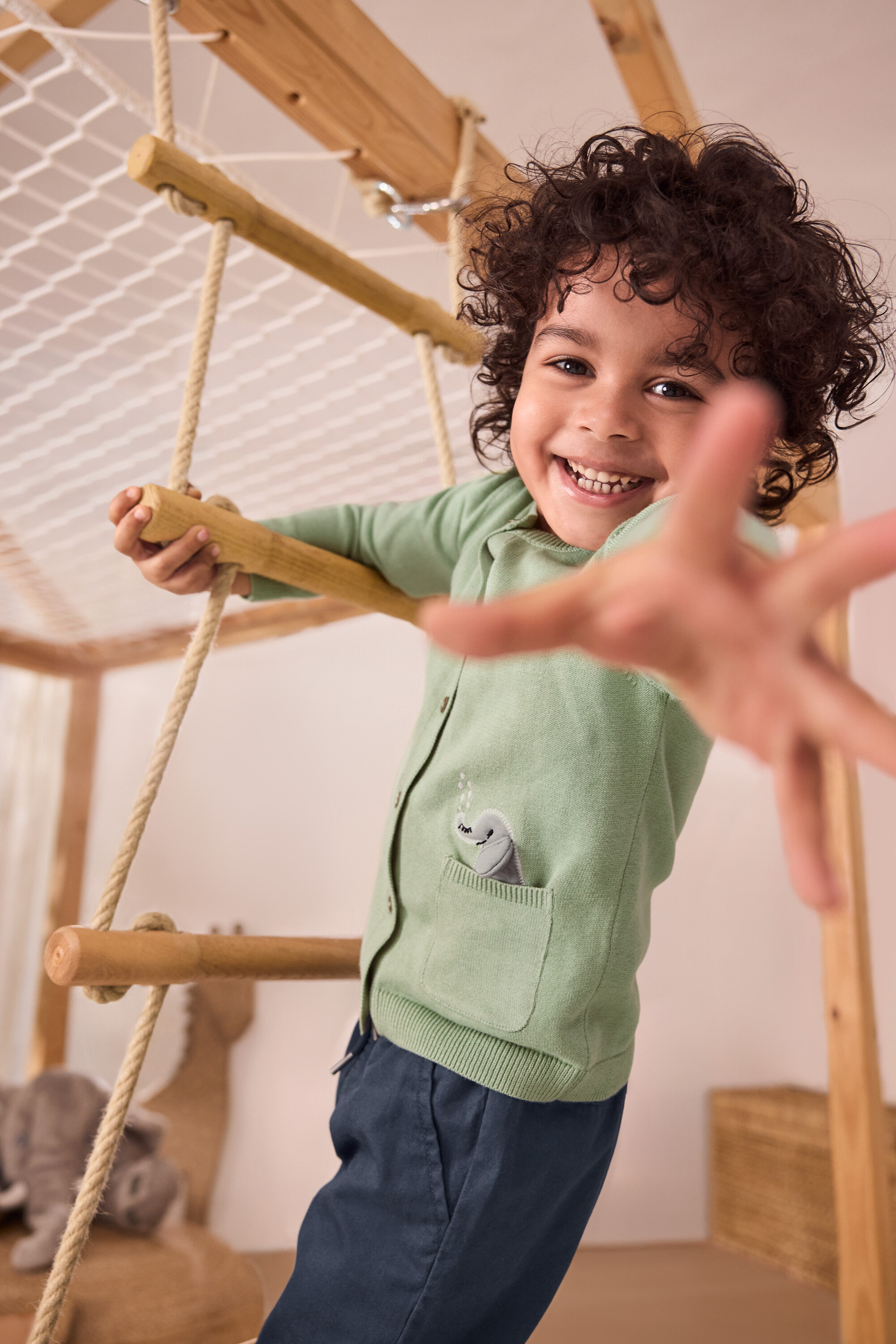 Un garçon souriant grimpe à une échelle de corde. Il porte une jaquette en maille pour bébé avec motif appliqué verte et un pantalon à enfiler pour enfant – Fit «
Anton
» bleu.
