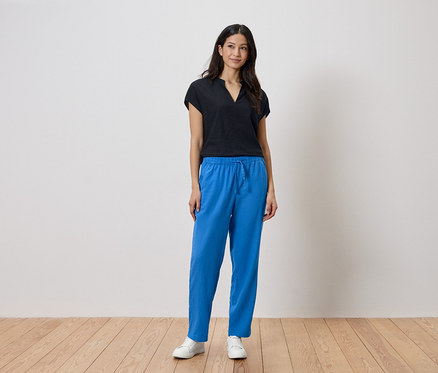 Femme debout devant un mur, portant un haut noir et un pantalon bleu.