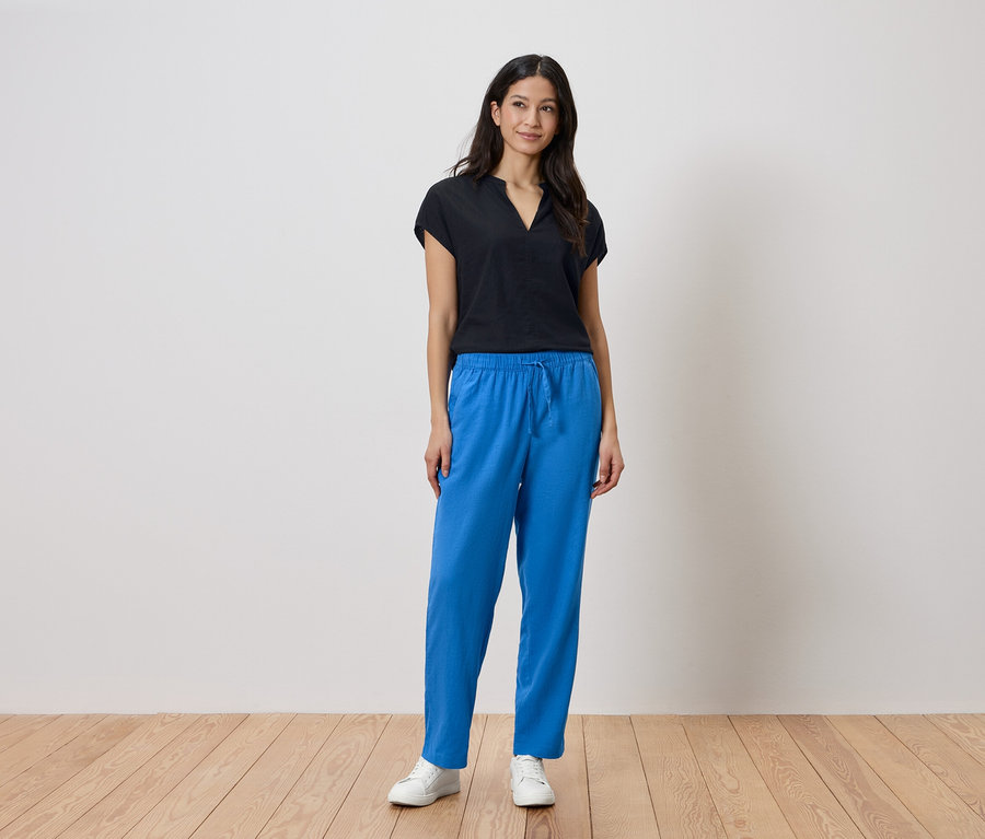 Femme debout devant un mur, portant un haut noir et un pantalon bleu.
