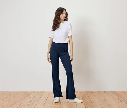 Femme debout en t-shirt blanc avec des cœurs et un pantalon évasé bleu devant un mur blanc.