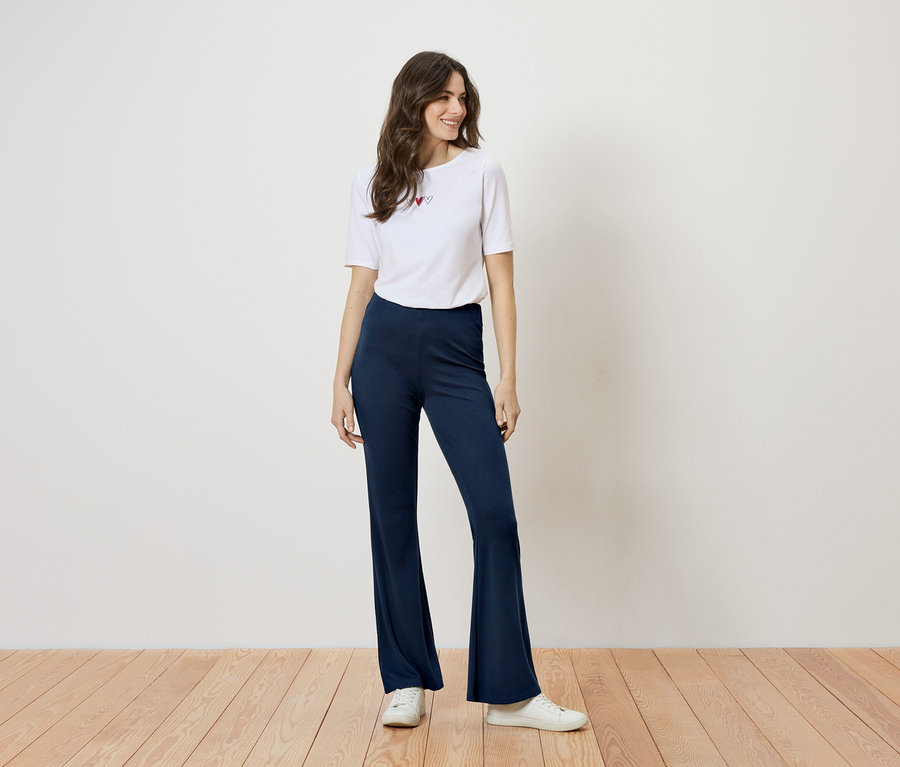 Femme debout en t-shirt blanc avec des cœurs et un pantalon évasé bleu devant un mur blanc.