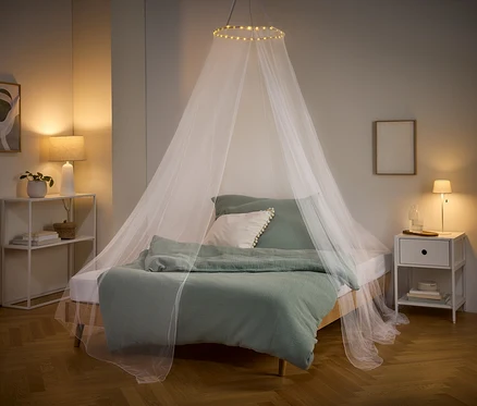 Un lit avec du linge de lit vert et une moustiquaire blanche avec des lumières suspendues au plafond. Une table de chevet et une étagère se trouvent sur les côtés.