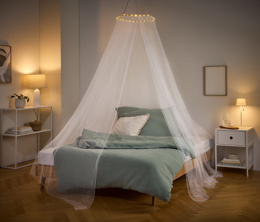 Un lit avec du linge de lit vert et une moustiquaire blanche avec des lumières suspendues au plafond. Une table de chevet et une étagère se trouvent sur les côtés.