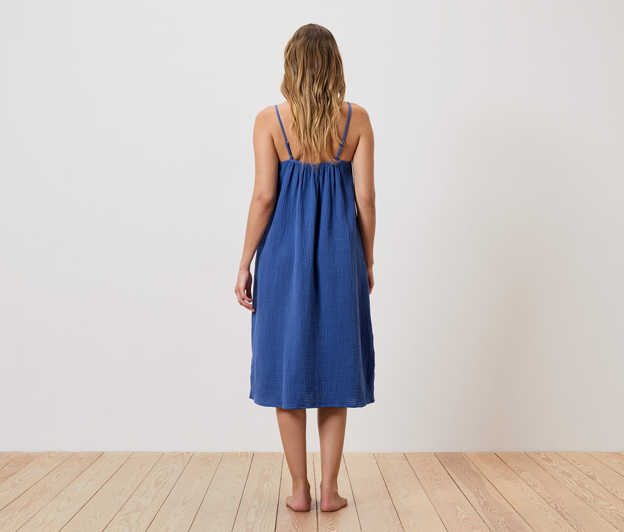 Une femme aux cheveux blonds et ondulés se tient dos à nous, portant une robe bleue à bretelles.