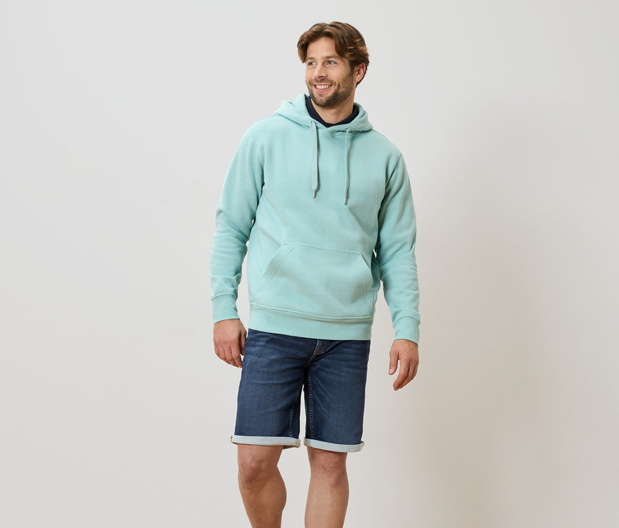 Un homme debout porte un sweat à capuche turquoise clair et un short en jean bleu.