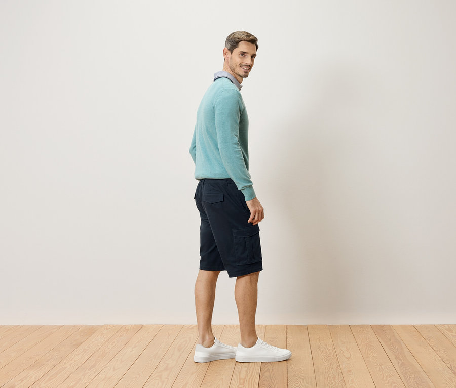 Un homme se tient de côté, regardant par-dessus son épaule, portant un pull bleu clair, un short cargo court foncé et des baskets blanches.