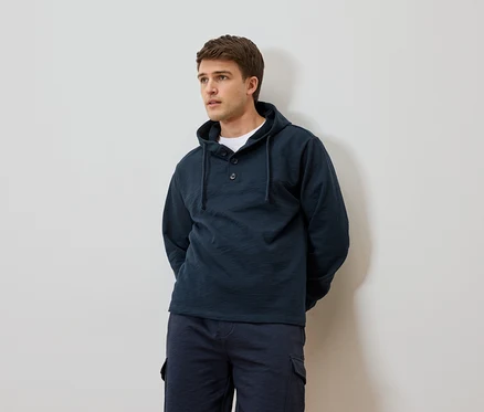 Homme portant un sweat à capuche bleu marine et un short devant un mur blanc.