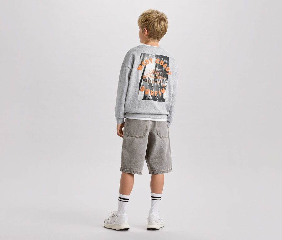 Garçon vu de dos, portant un sweat-shirt gris imprimé, un short et des chaussures de sport.