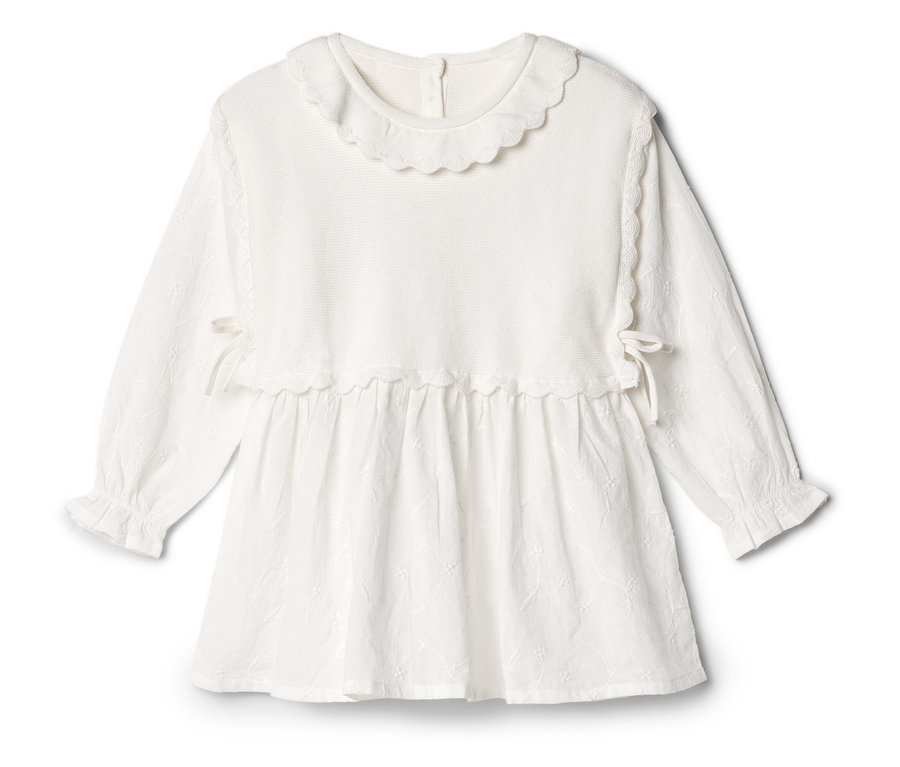 Une robe blanche à manches longues avec un col à volants repose sur un fond blanc.