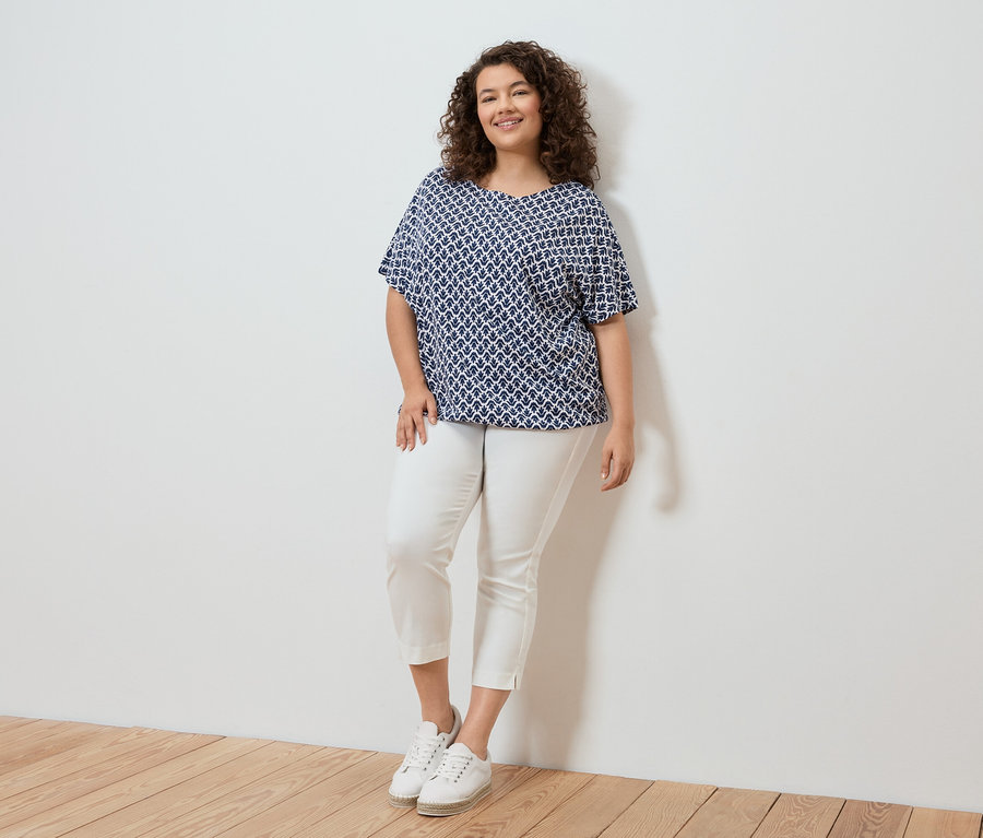 Femme aux cheveux bouclés portant un chemisier à motifs bleu et blanc, un pantalon blanc et des baskets blanches debout devant un mur blanc.