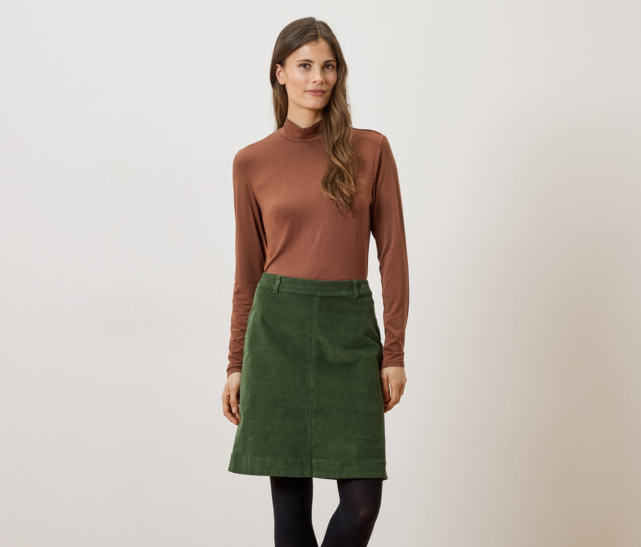 Femme portant un pull à col roulé marron et une jupe en velours côtelé verte, avec des collants noirs sur un fond clair.