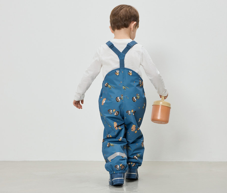 Enfant vu de dos portant une salopette de pluie bleue à motifs d'animaux et un haut blanc, tenant une boîte à la main.
