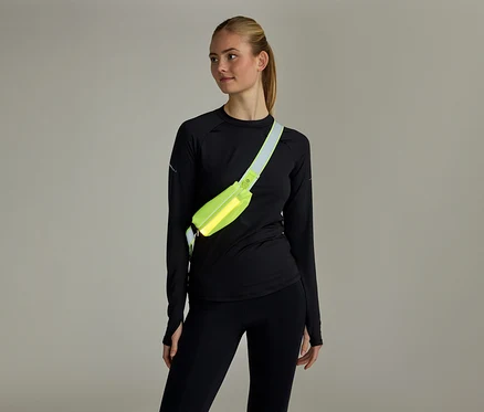 Femme aux cheveux attachés, vêtue d'un haut noir à manches longues et d'un legging noir, portant un sac banane jaune fluo en bandoulière.