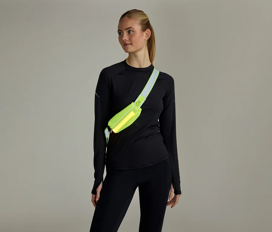 Femme aux cheveux attachés, vêtue d'un haut noir à manches longues et d'un legging noir, portant un sac banane jaune fluo en bandoulière.