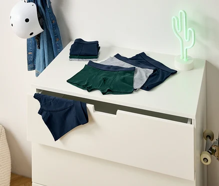 Des boxers verts, gris et bleus sont posés sur une commode. Un boxer bleu dépasse d'un tiroir. Un skateboard est appuyé contre le mur.
