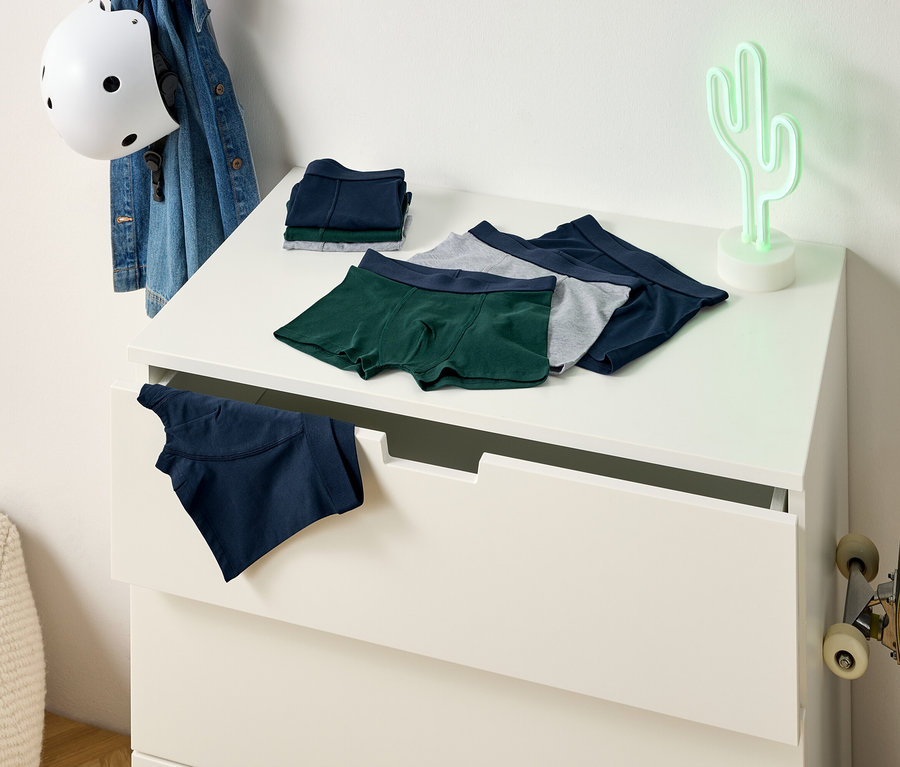 Des boxers verts, gris et bleus sont posés sur une commode. Un boxer bleu dépasse d'un tiroir. Un skateboard est appuyé contre le mur.