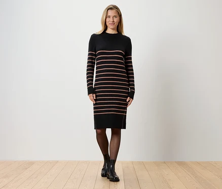 Une femme blonde se tient devant un mur blanc, portant une robe en tricot noire rayée, des collants noirs et des bottes noires.