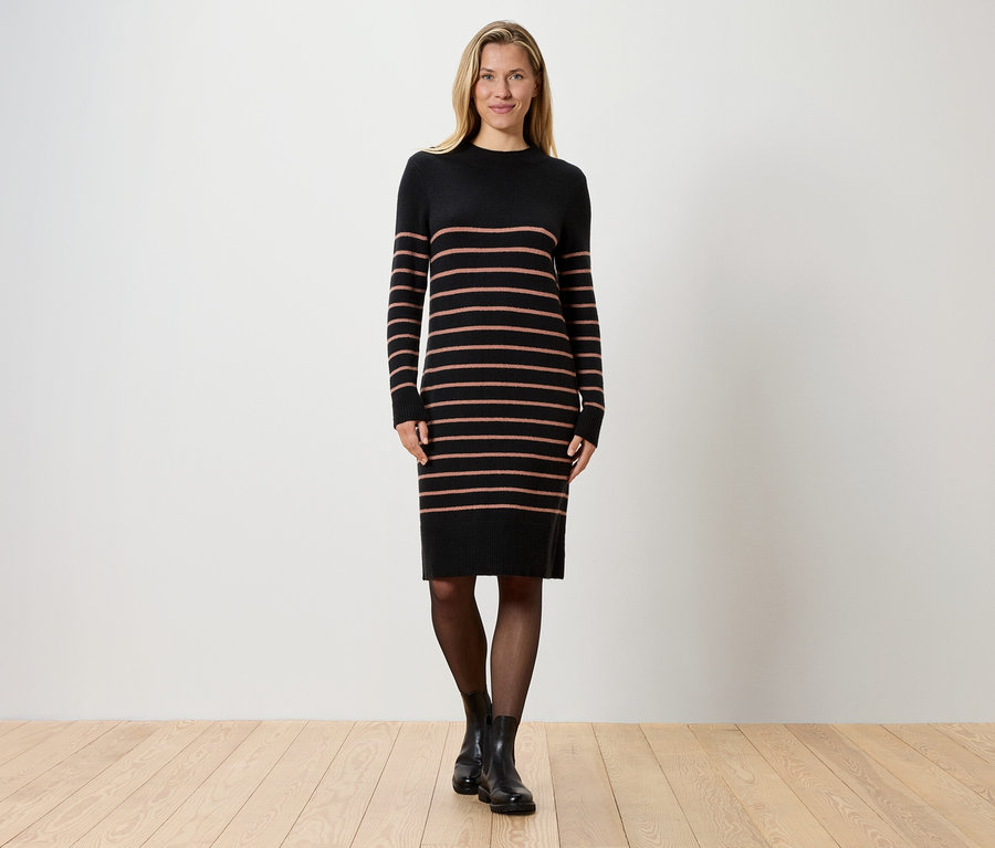 Une femme blonde se tient devant un mur blanc, portant une robe en tricot noire rayée, des collants noirs et des bottes noires.