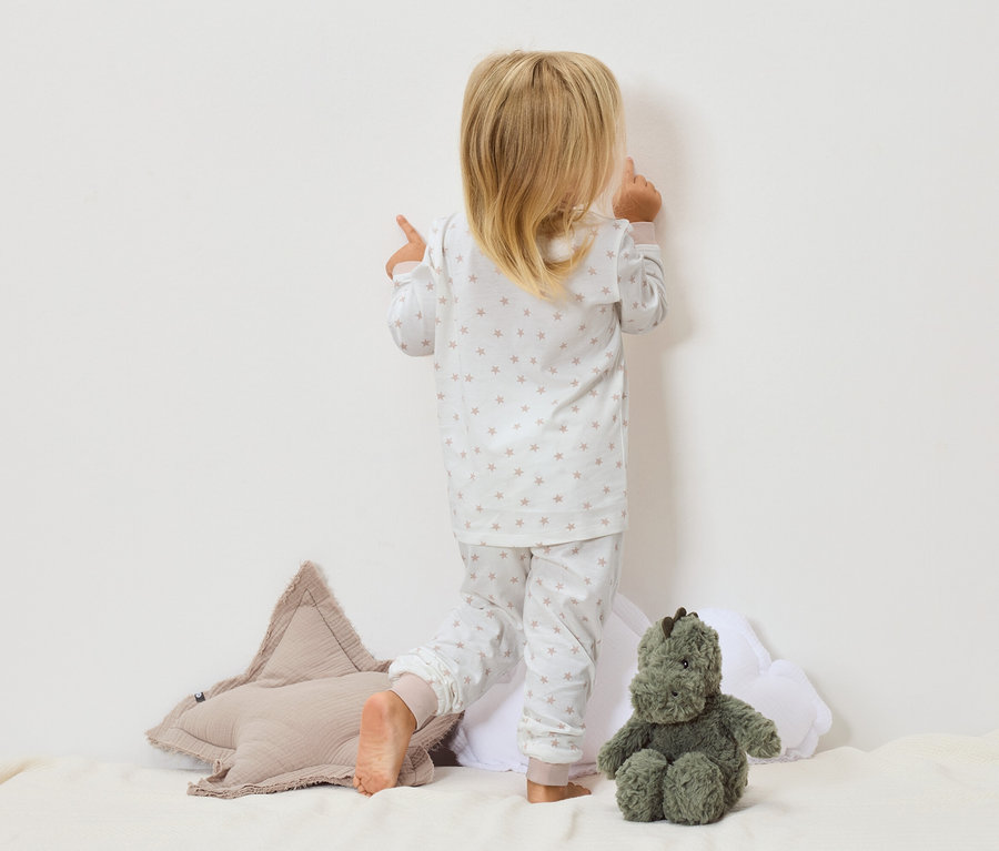 Un enfant en pyjama étoilé se tient face au mur, à côté d'un coussin étoile et d'un jouet en peluche vert.