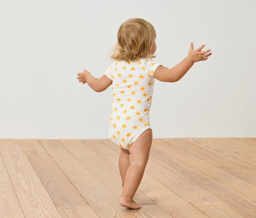 Un petit enfant aux cheveux blonds, portant un body à motifs de tournesols, se tient sur un plancher en bois.