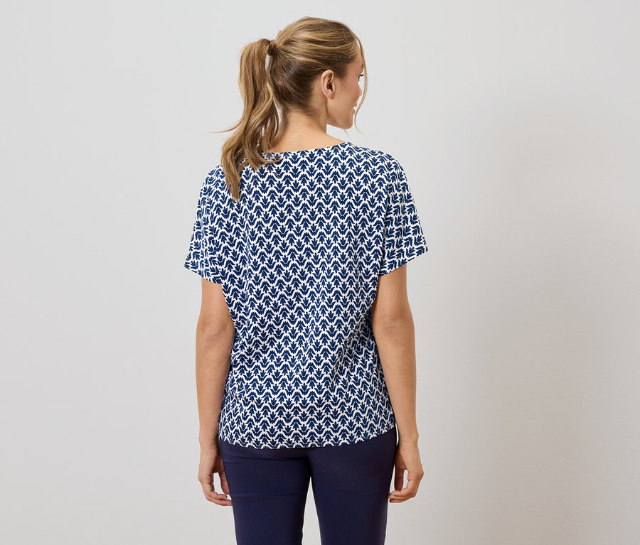 Vue de dos d'une femme avec une queue de cheval blonde portant un chemisier à manches courtes bleu marine et blanc à motifs de feuilles et un pantalon bleu marine.