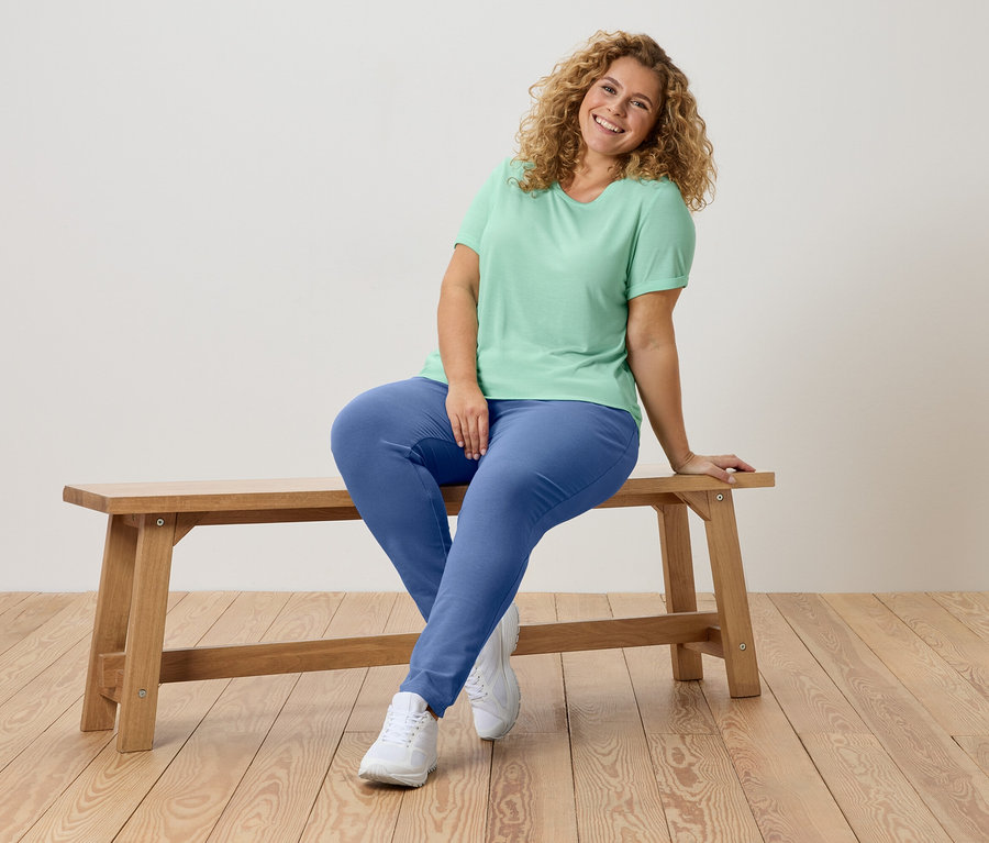 Une femme aux cheveux bouclés est assise sur un banc en bois, portant un t-shirt vert et un pantalon bleu.