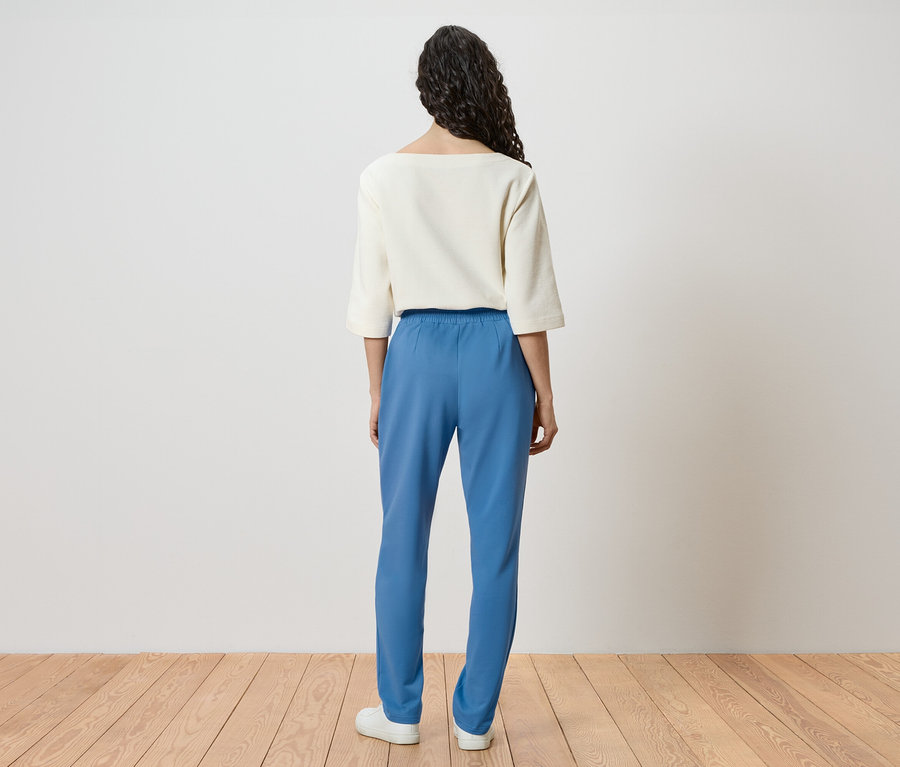 Vue arrière d'une femme aux cheveux bouclés portant un haut blanc, un pantalon bleu et des chaussures blanches.