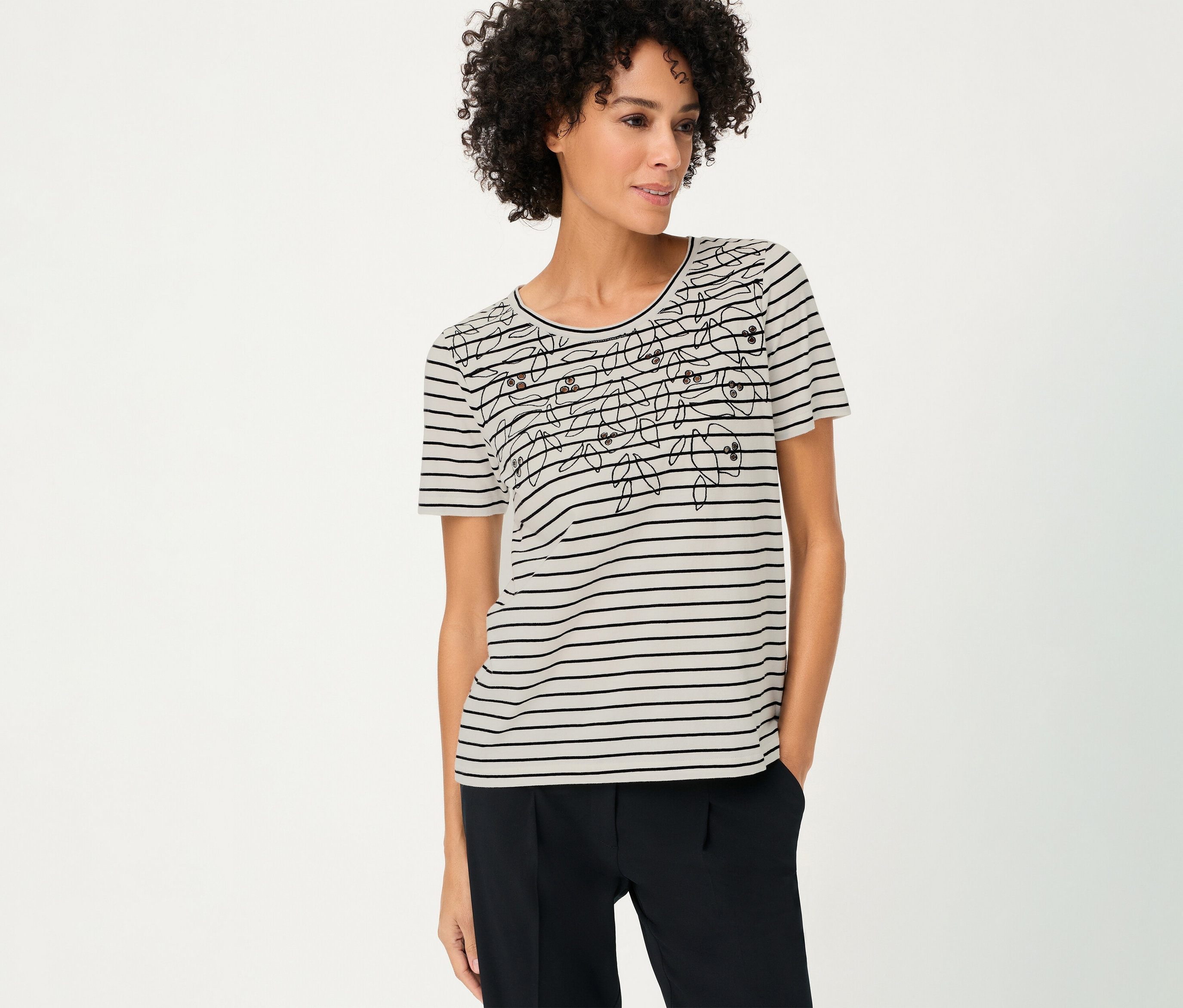 Une femme aux cheveux bouclés porte un T-shirt Olsen rayé avec un motif floral et un pantalon foncé.