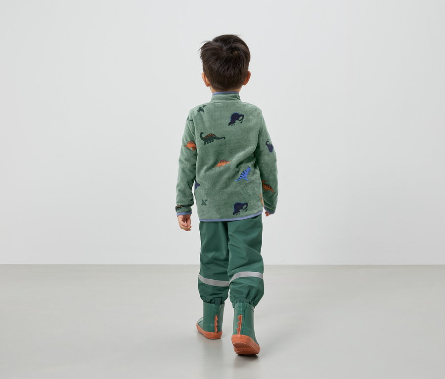 Un enfant vu de dos, porte un pull en polaire vert avec des dinosaures, un pantalon de pluie vert et des bottes en caoutchouc.