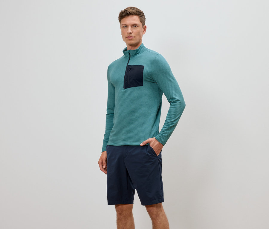 Homme portant un haut à manches longues turquoise avec fermeture éclair et un short bleu marine, une main dans la poche.