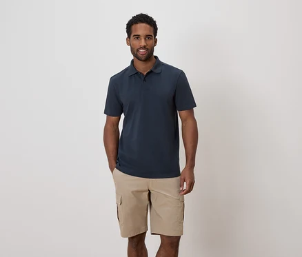 Un homme aux cheveux foncés et à la barbe porte un polo bleu marine et un short cargo kaki.