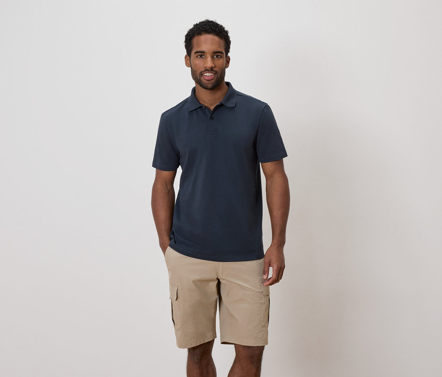 Un homme aux cheveux foncés et à la barbe porte un polo bleu marine et un short cargo kaki.