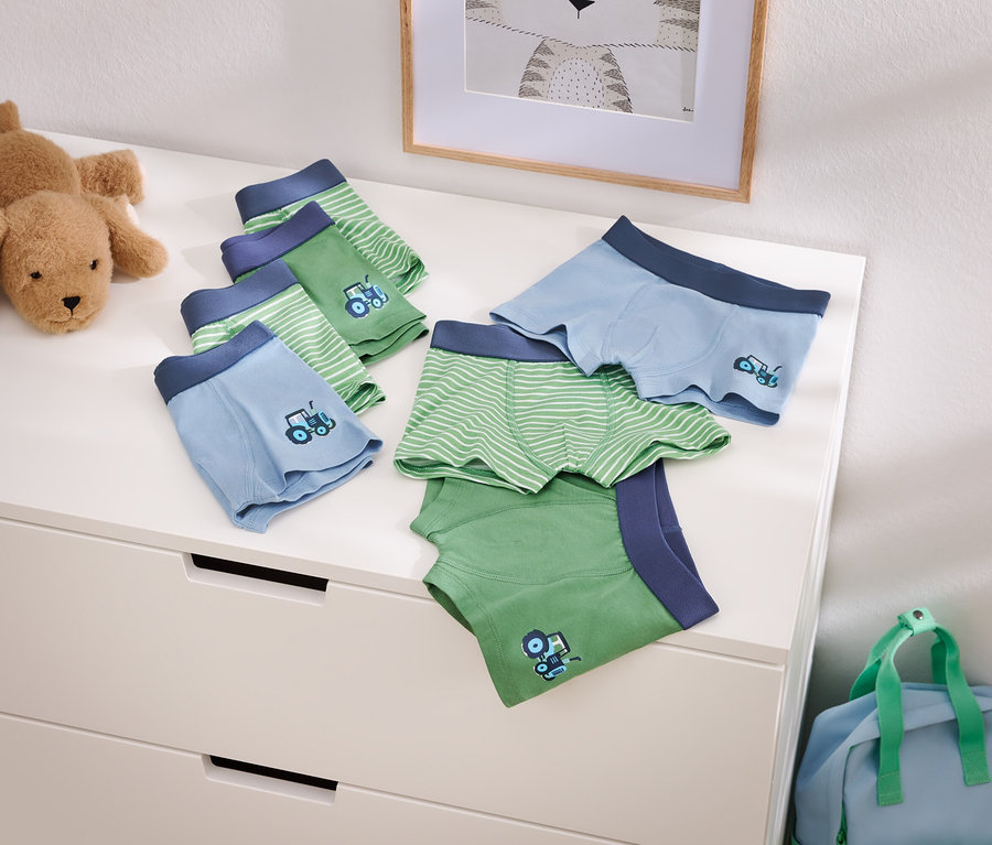 Photo de boxers pour garçons bleu clair et vert avec un motif de tracteur sur une commode, à côté d'un chien en peluche et d'un sac de voyage bleu.