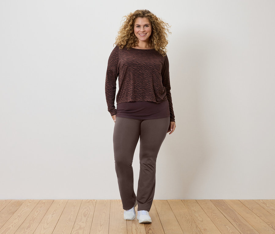 Une femme aux cheveux bouclés porte un haut à manches longues marron à motifs, un haut plus foncé et un pantalon marron, ainsi que des baskets blanches.