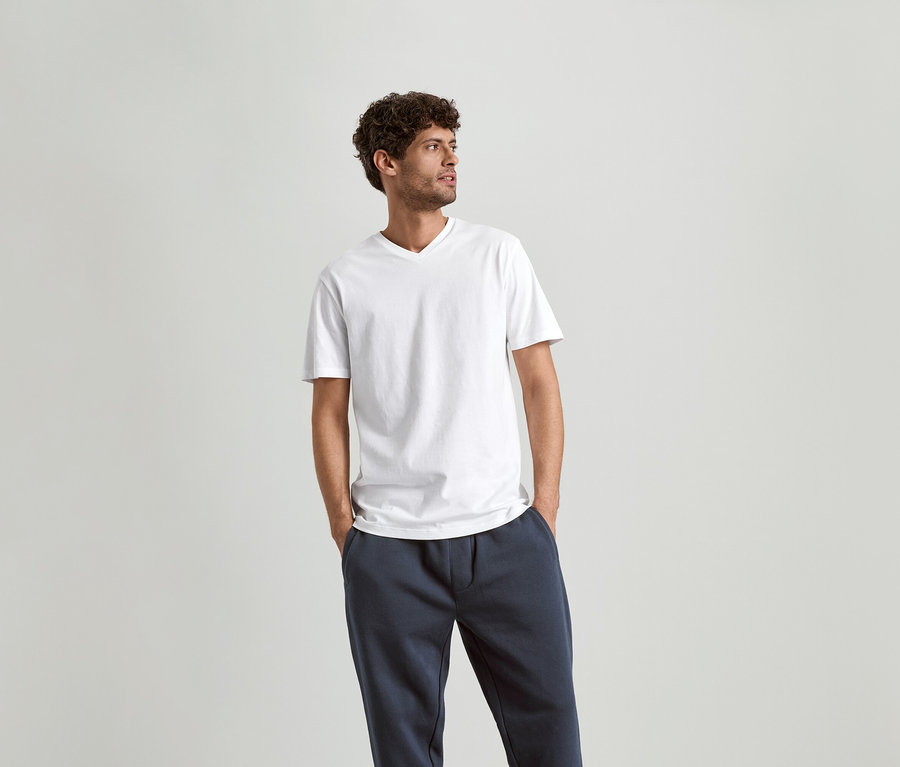 Homme aux cheveux bouclés portant un t-shirt blanc à col en V et un pantalon de jogging bleu marine.