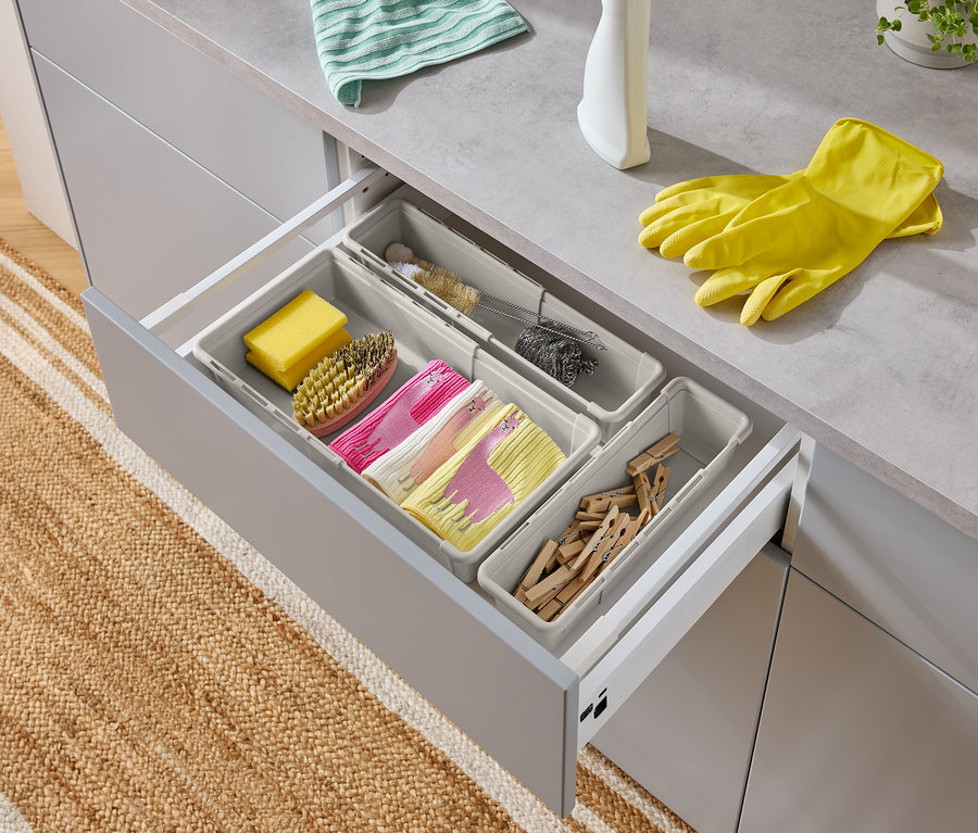 Tiroir ouvert avec des ustensiles de lavage : éponge, brosse, torchons avec motif lama, goupillon, tampon à récurer, pinces à linge et gants jaunes.