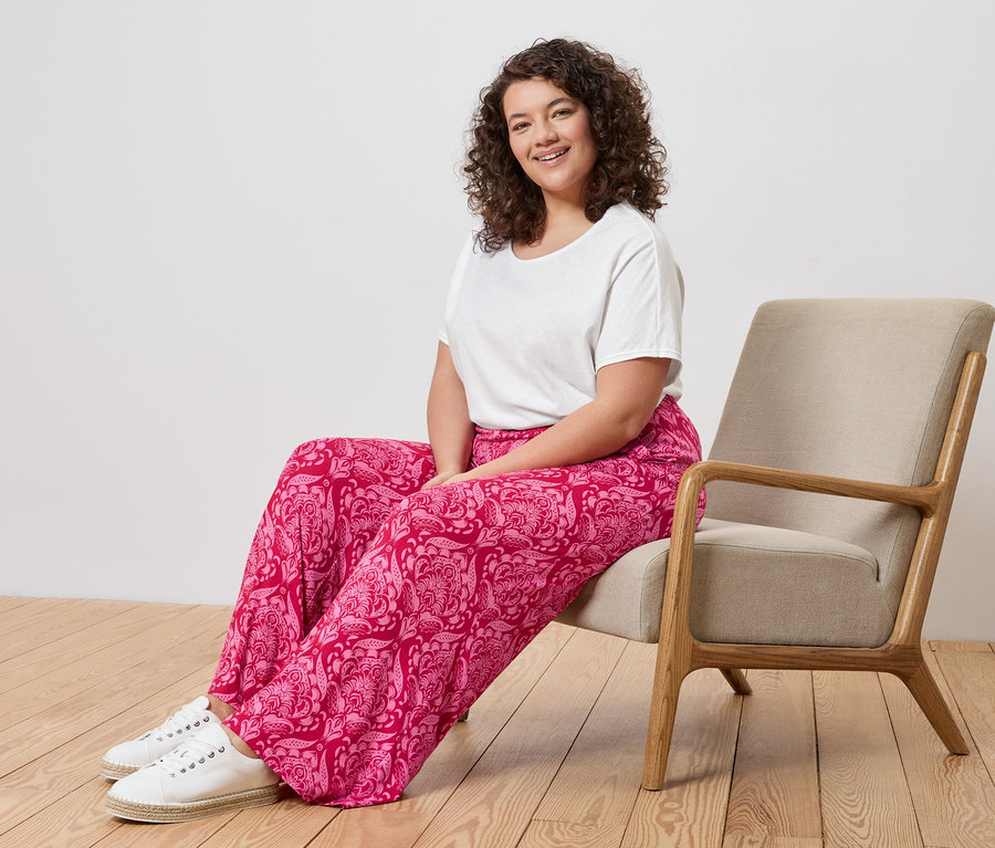 Une femme aux cheveux bouclés est assise dans un fauteuil beige, portant un t-shirt blanc, un pantalon rose à motifs cachemire et des baskets blanches.