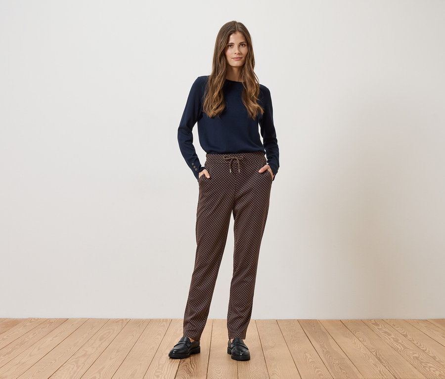 Une femme se tient devant un mur blanc, portant un pull bleu marine, un pantalon à motifs et des chaussures noires.