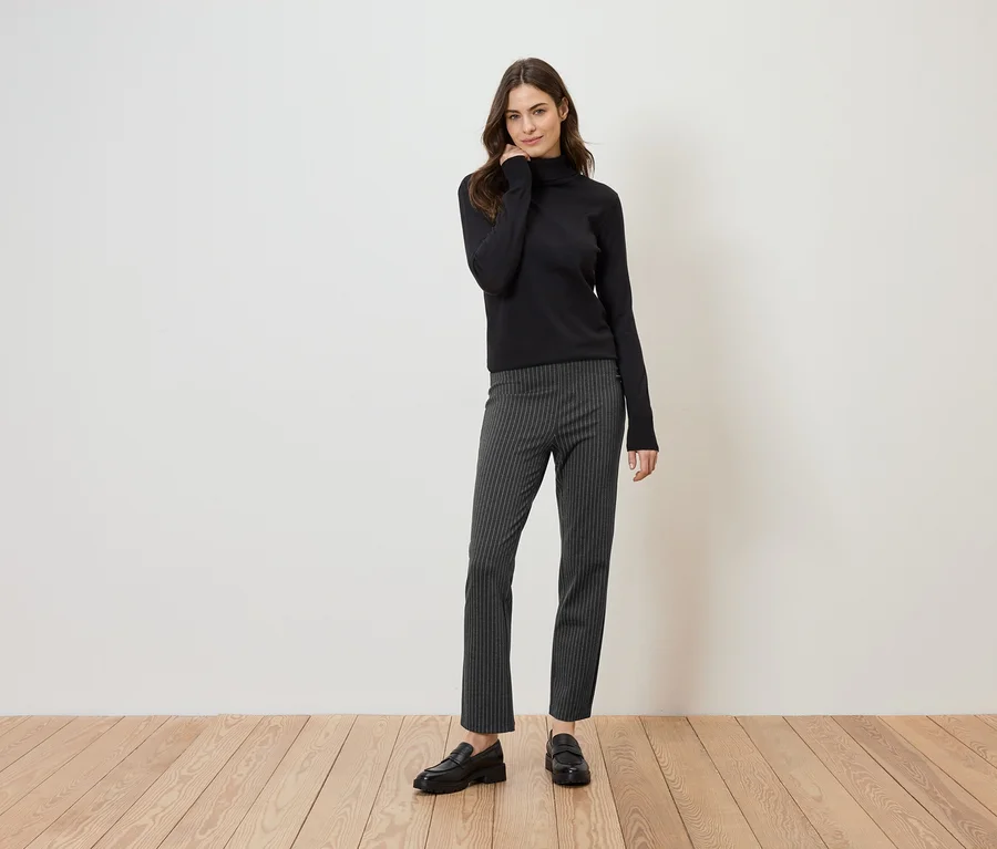 Une femme portant un pull à col roulé noir, un pantalon rayé et des mocassins noirs se tient sur un plancher en bois devant un mur blanc.