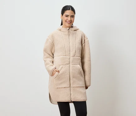 Une femme souriante portant un long manteau en fausse fourrure beige à capuche et des leggings noirs.